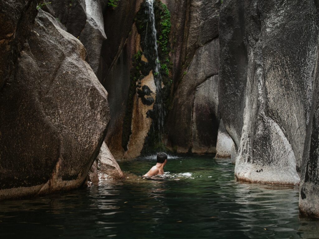 Tranquil natural pool nestled between giant granite boulders, featuring a swimmer relaxing under a cascading waterfall in a lush canyon setting
