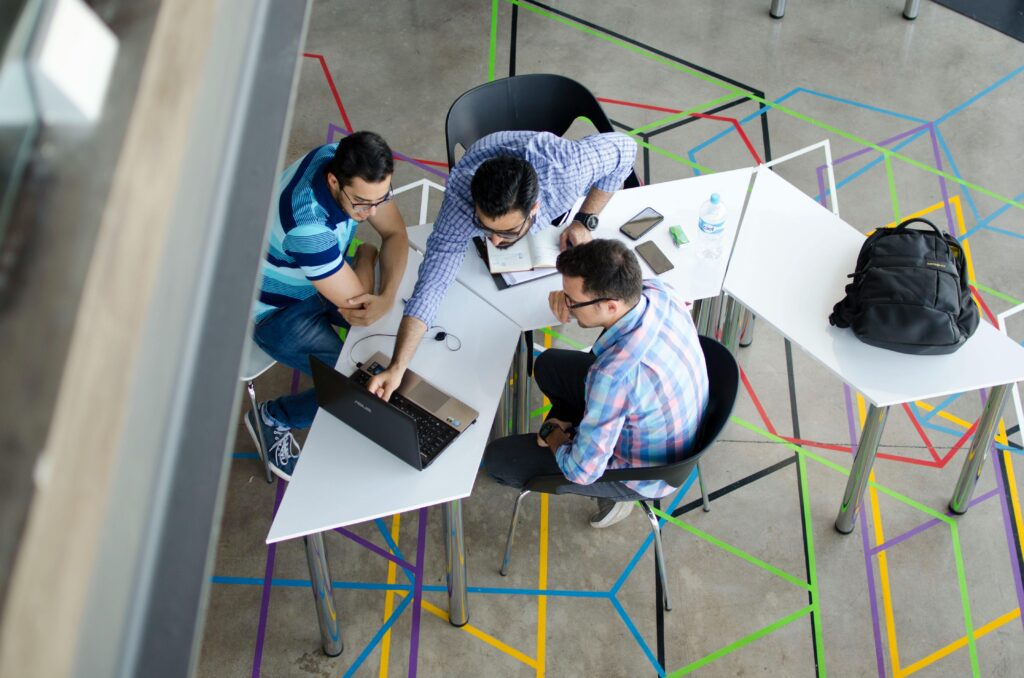 Three colleagues collaborate on a project, gathered around a laptop in a modern office space with colorful geometric floor designs.
