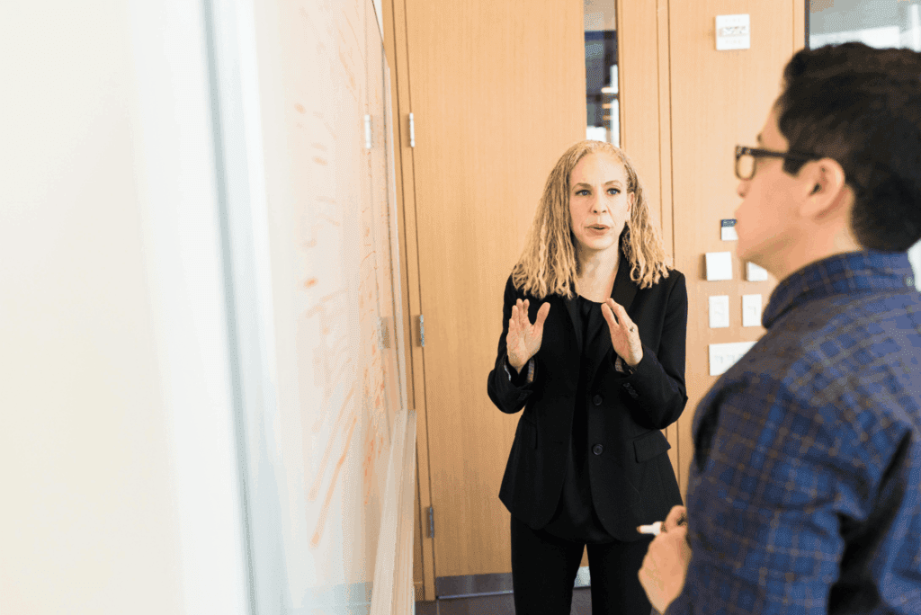 A businesswoman explains a concept to a colleague while standing at a whiteboard during a workplace meeting