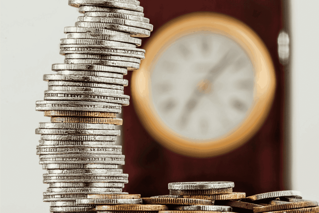 A stack of coins is shown in sharp focus with a blurred clock in the background, symbolizing the concept of time and money