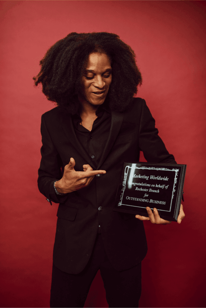 A man in a black suit smiles while proudly pointing to his business award plaque