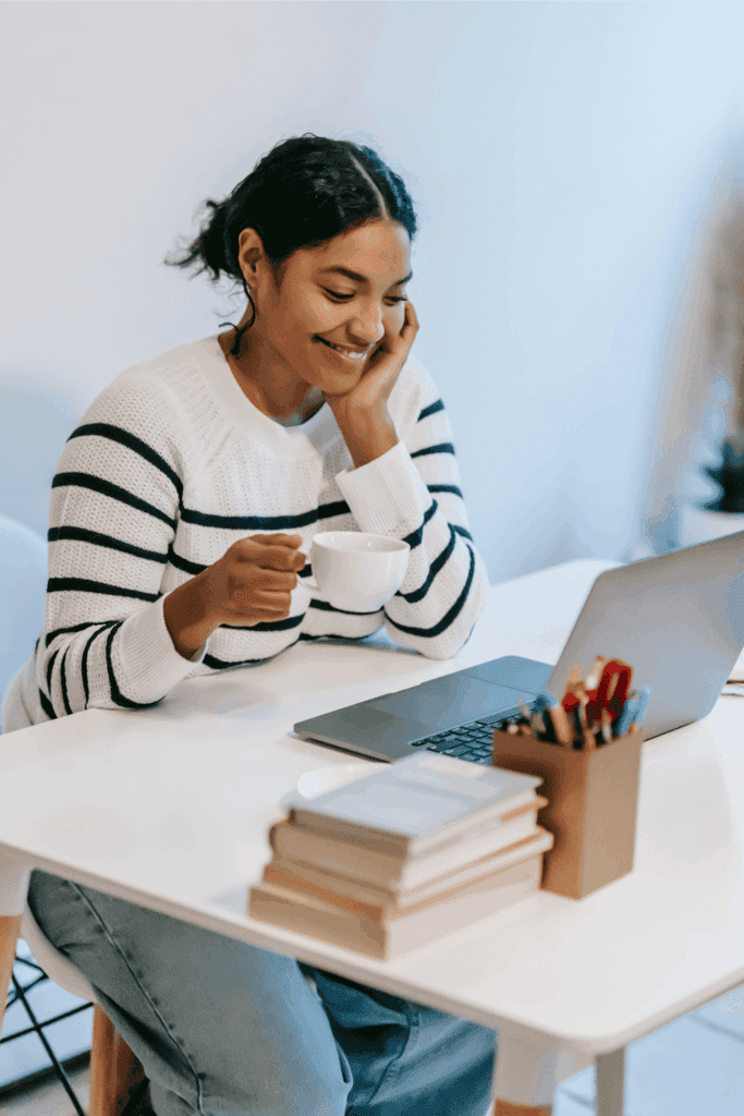 Woman enjoying coffee while working on her laptop at a bright, organized desk, creating a relaxed and productive work-from-home atmosphere