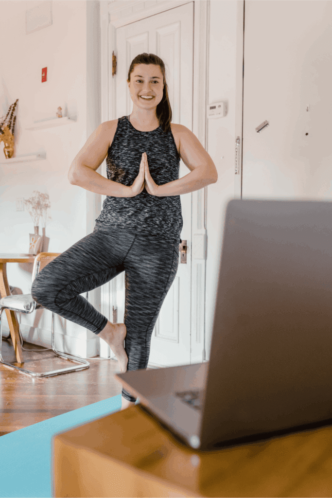 Woman practicing yoga in tree pose while following an online class on her laptop, promoting health and wellness at home