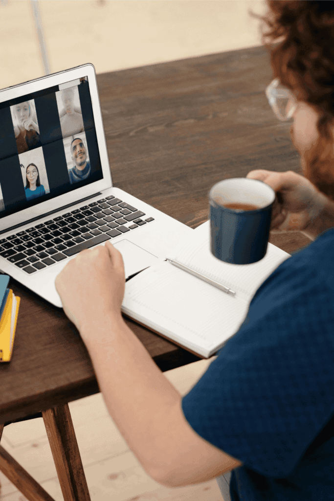 Person participating in a virtual meeting on a laptop while taking notes and drinking coffee, representing remote teamwork and online collaboration