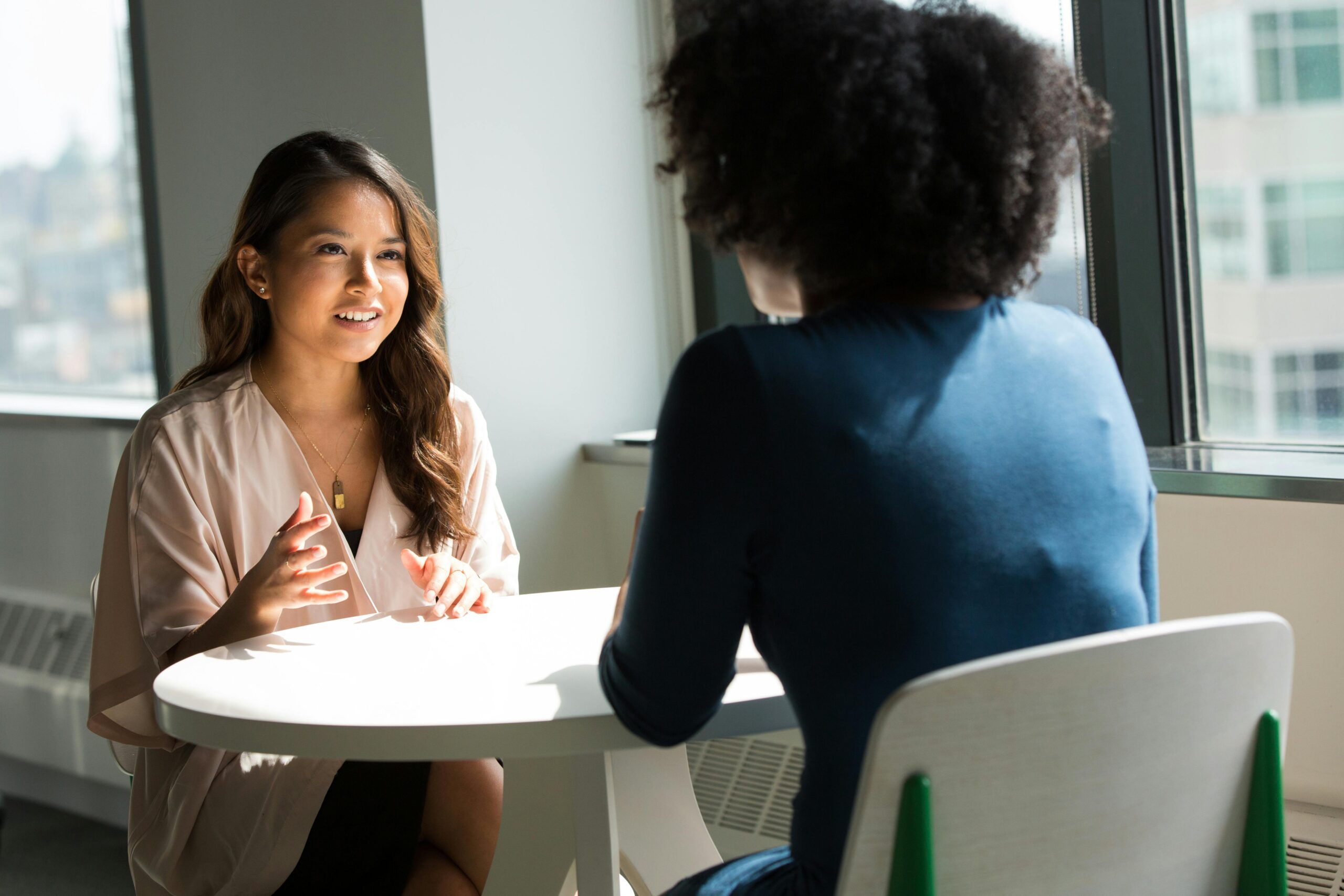 Two women sit at a small table having a conversation in a bright office