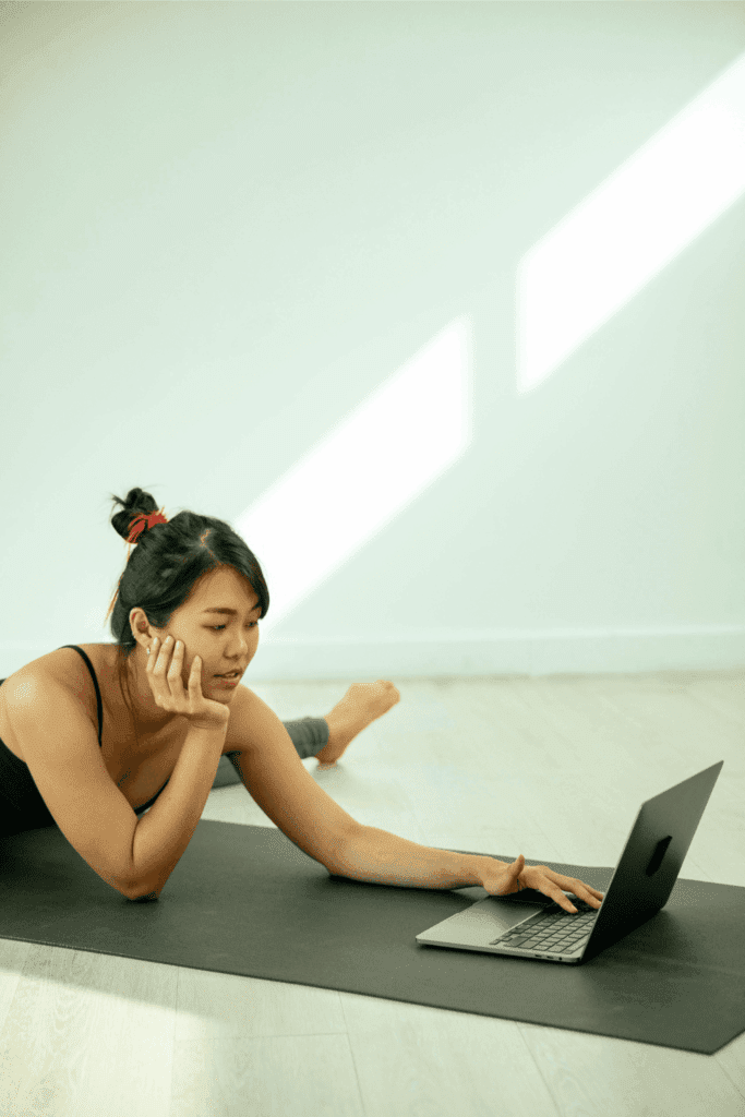 A woman stretches on a yoga mat while watching something on her laptop.
