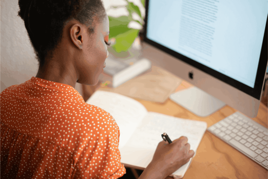 Professional woman taking notes while working on a computer in a modern home office workspace.
