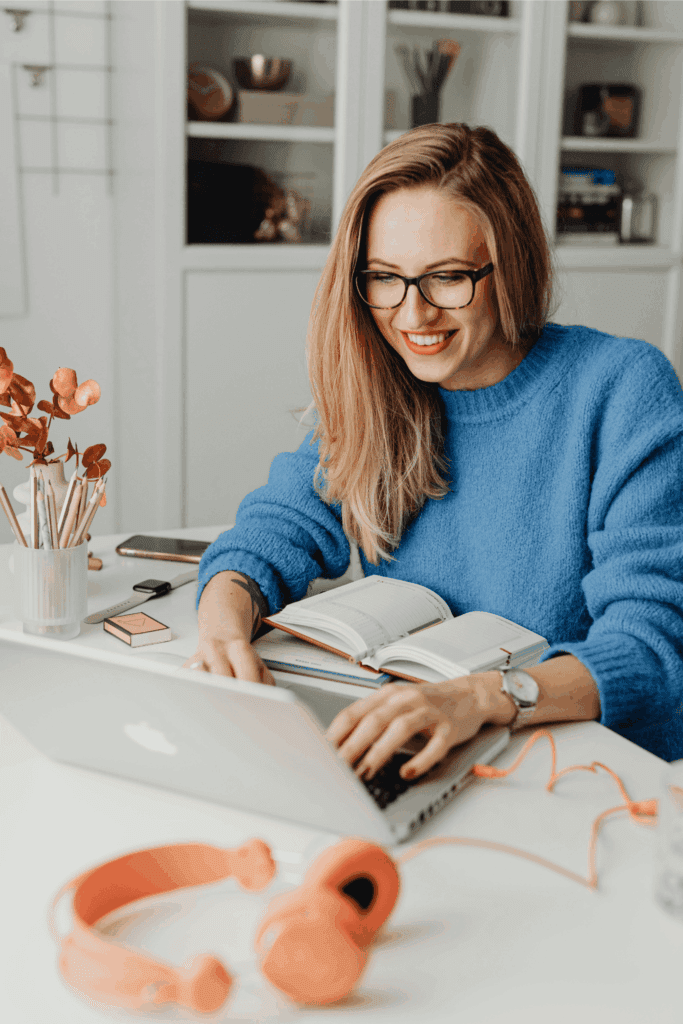 Smiling woman in glasses working on a laptop with an open notebook – remote work, productivity, and home office concept.
