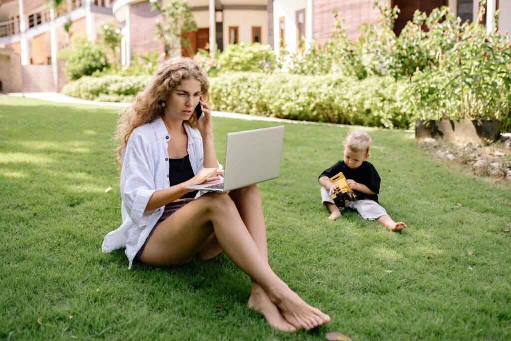 Woman working remotely on a laptop in the garden while her child plays nearby – work-life balance and remote work lifestyle concept.
