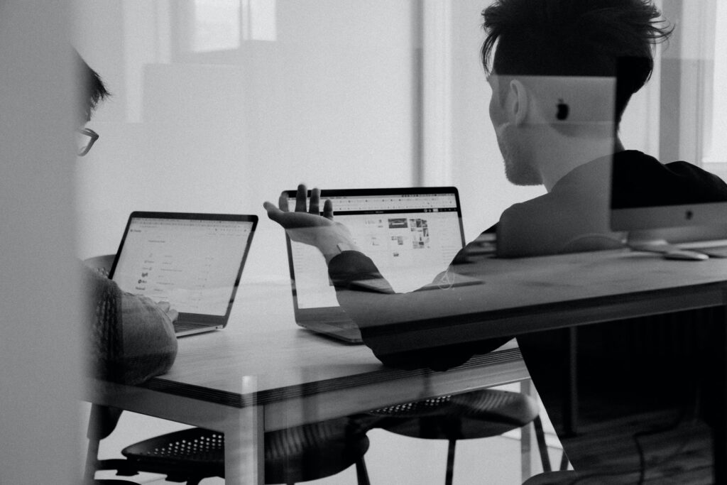 two employees sitting at a desk in a meeting room with their laptops giving feedback