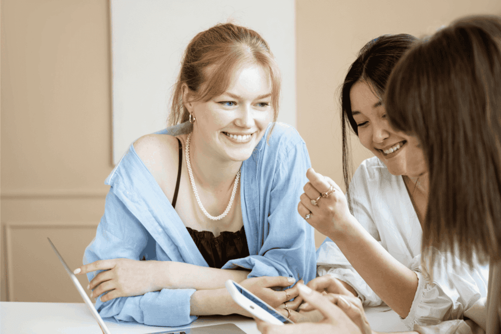 Group of young women smiling and collaborating while looking at a phone – teamwork, friendship, and modern communication concept.
