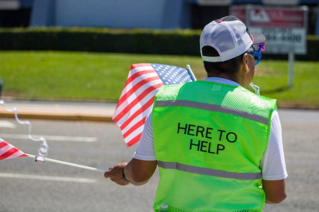 an employee volunteering on the road with a flag and a vest that says here to help