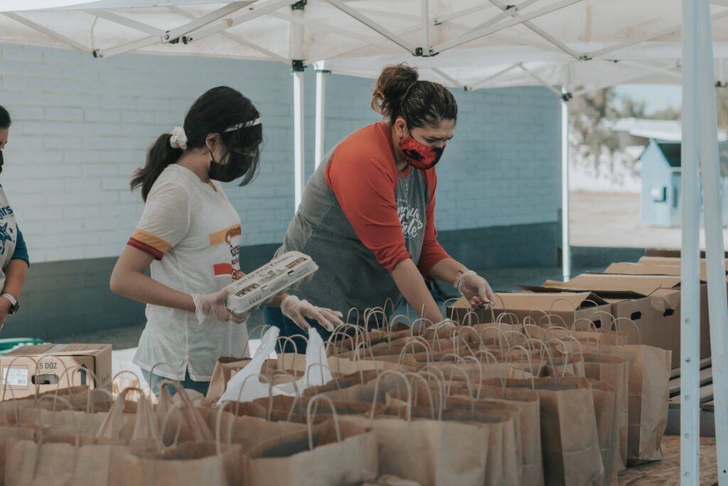 two employees volunteering and filling paper bags with groceries under a tent