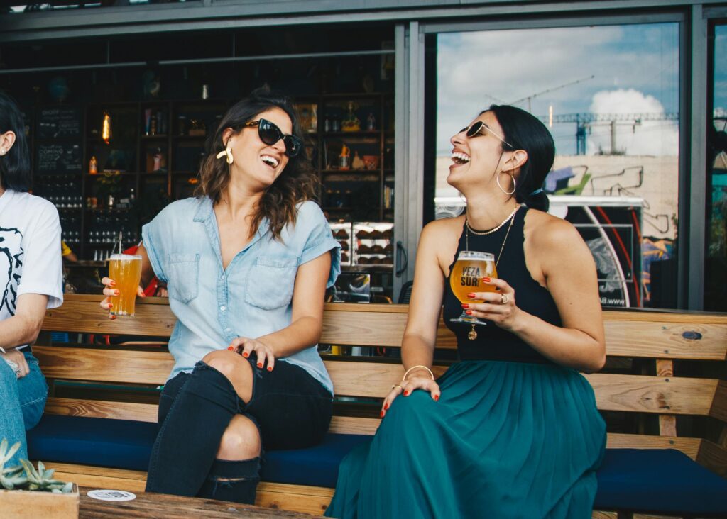 Two women laughing and enjoying drinks at an outdoor bar – friendship, social life, and casual fun concept