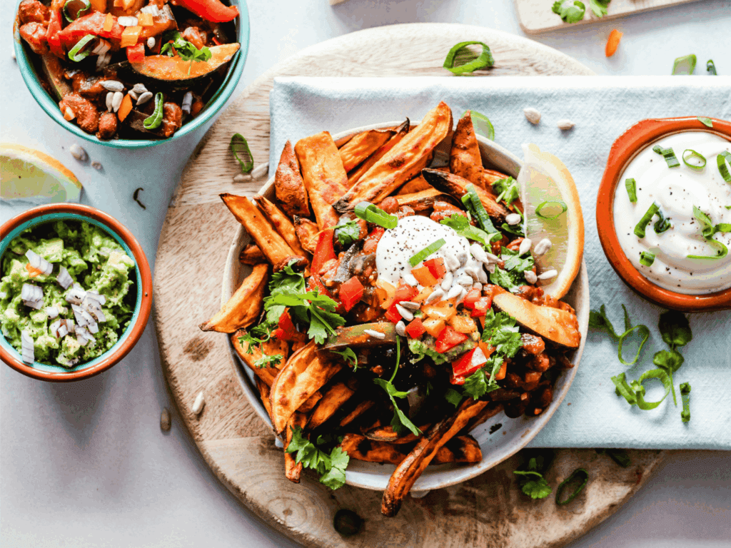 A colorful plate of sweet potato fries topped with fresh vegetables, herbs, and sour cream is served with sides of guacamole, salsa, and dipping sauce for a vibrant, healthy meal.
