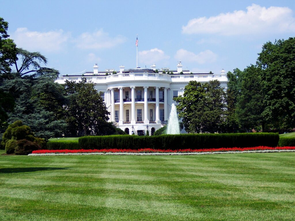 The White House in Washington, D.C., with its iconic white columns, central fountain, and well-manicured lawn framed by trees and flowerbeds.
