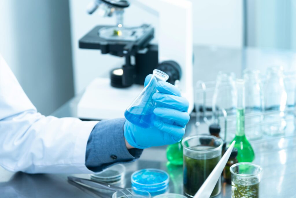 A scientist wearing gloves holds a flask of blue liquid in a laboratory, surrounded by glassware, samples, and a microscope for research and experiments.
