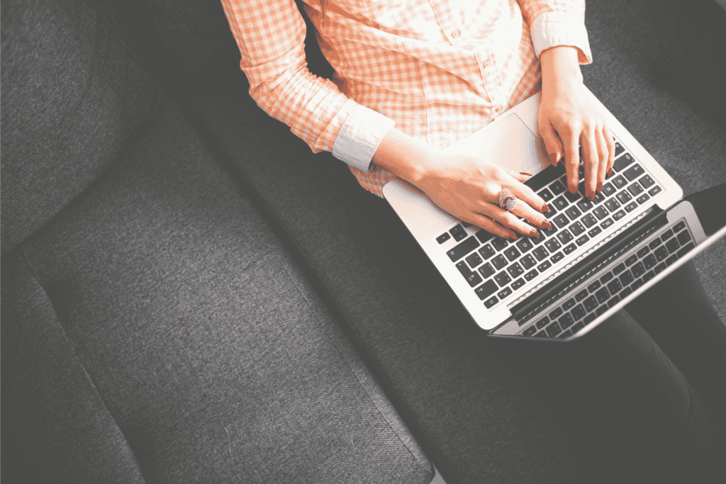 A woman types on a laptop while sitting on a gray couch.
