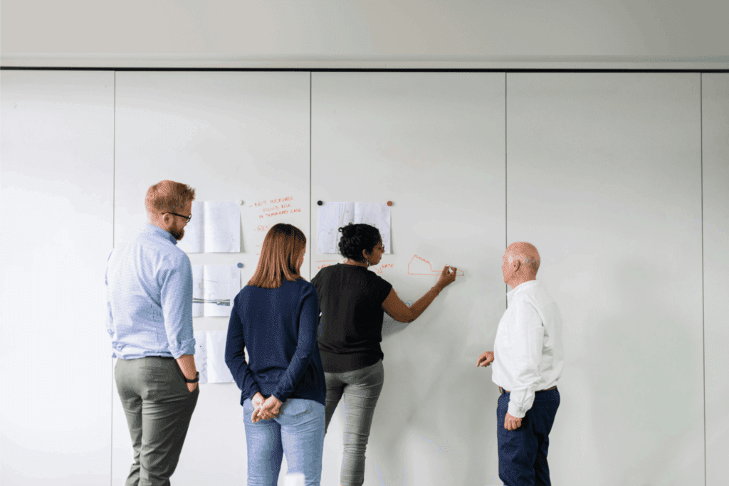 A diverse team collaborates by sharing ideas and problem-solving together on a whiteboard.