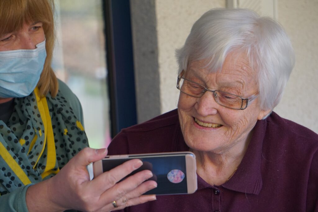 a woman volunterring at a nursing home showing a smiling elderly woman something on her phone