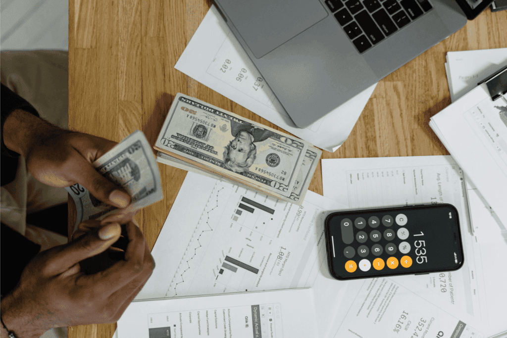 A person calculates finances with cash, reports, and a calculator spread out on a desk.