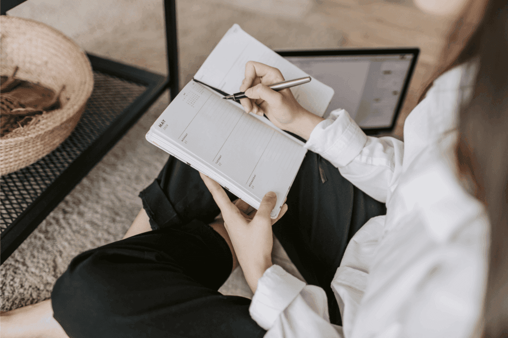 A person writes plans in a notebook planner while sitting with a laptop nearby.