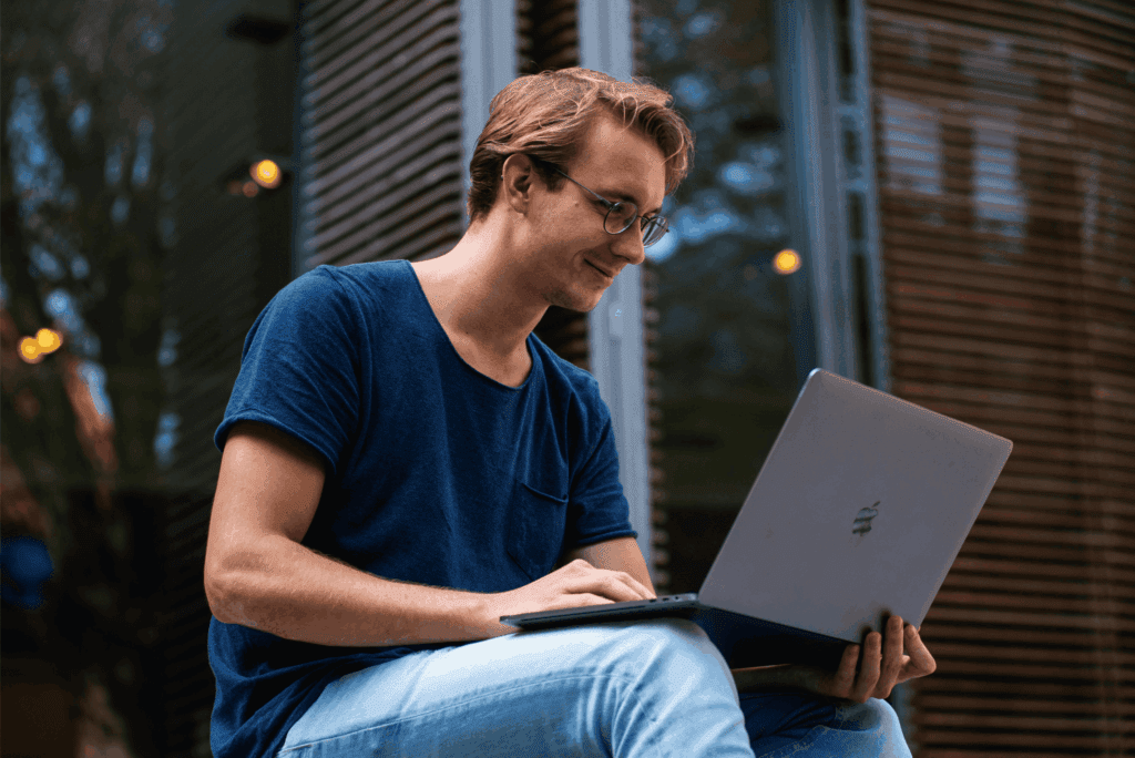 Man sitting outdoors smiling while working on a laptop.