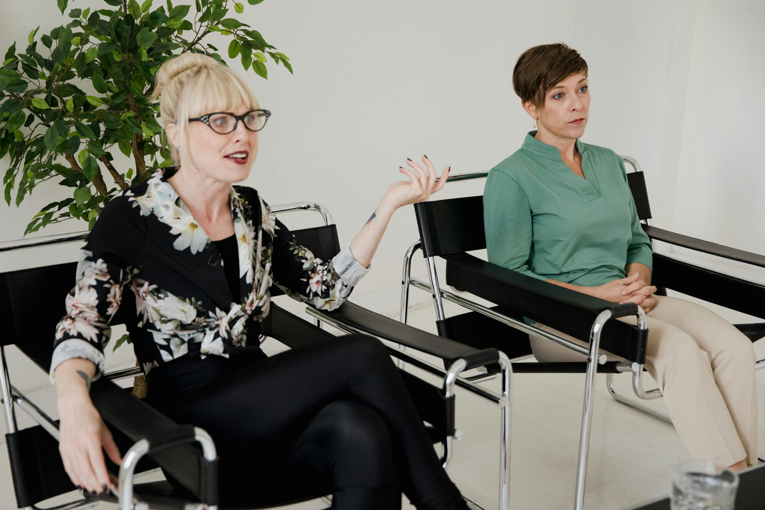 Two women sit in chairs during a discussion, one speaking passionately while the other listens attentively.