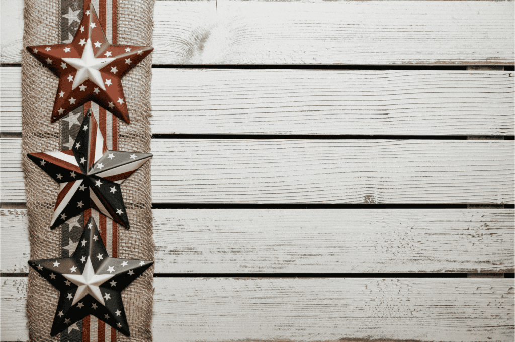 Three patriotic stars with American flag patterns hang on burlap beside white wooden planks.