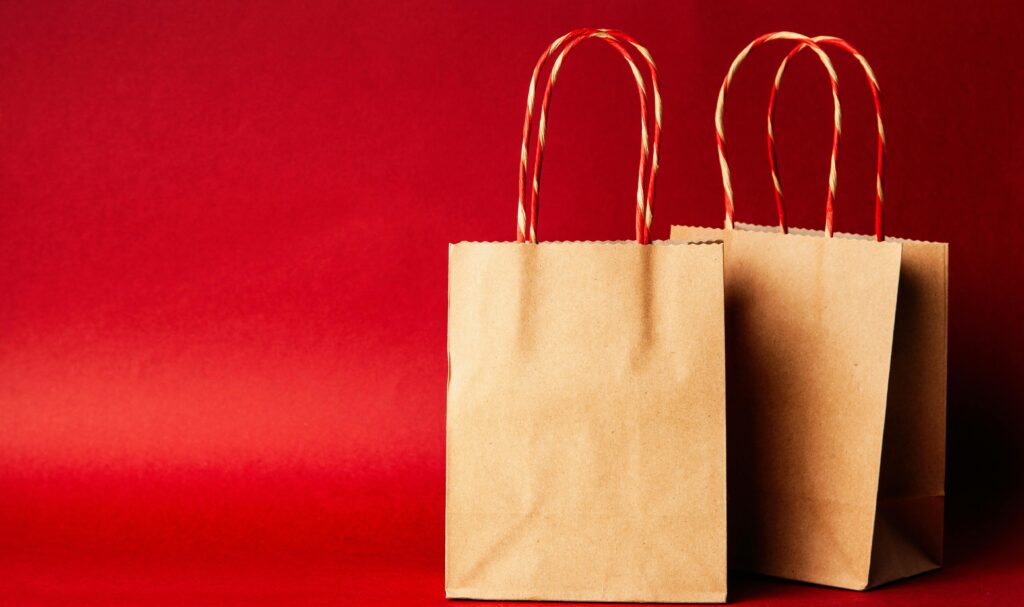 Two brown paper gift bags with red twisted handles stand against a bright red background.
