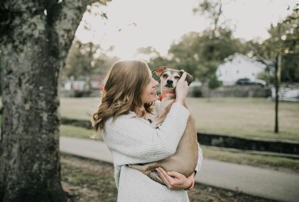 A woman in a cozy sweater lovingly holds her dog in her arms while smiling at it in a peaceful outdoor park setting.
