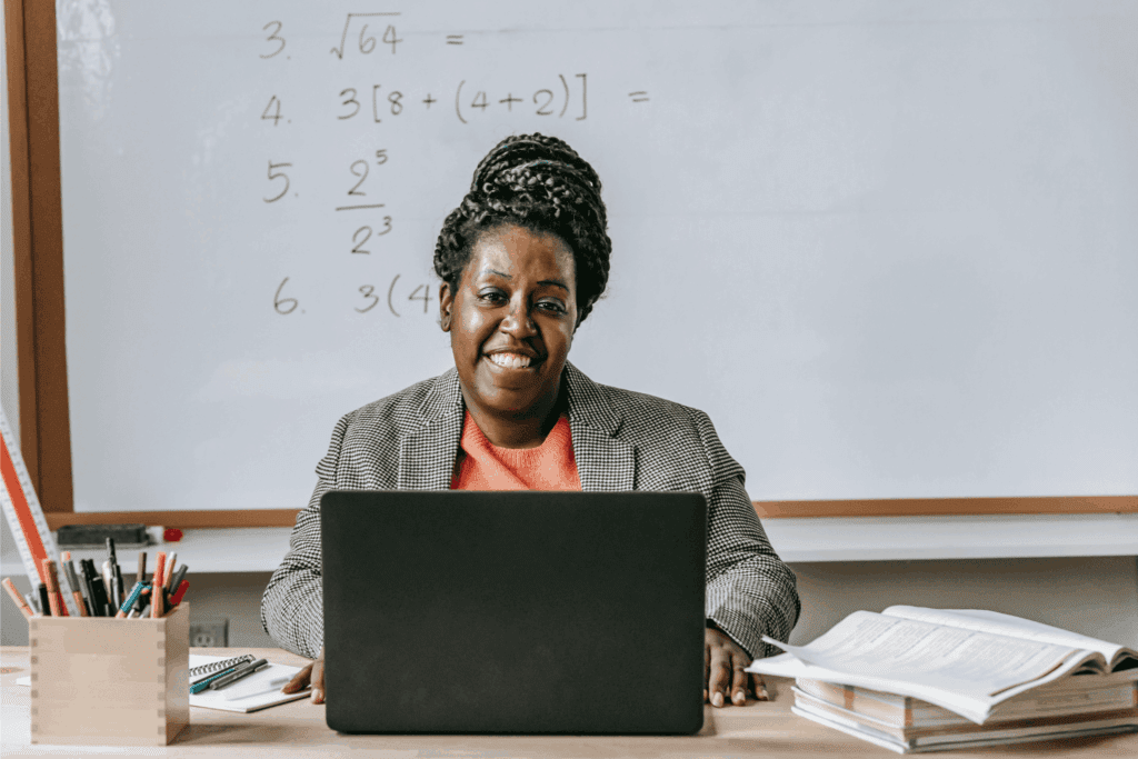 A smiling teacher sits at a desk with a laptop, open books, and pens, in front of a whiteboard filled with math problems.
