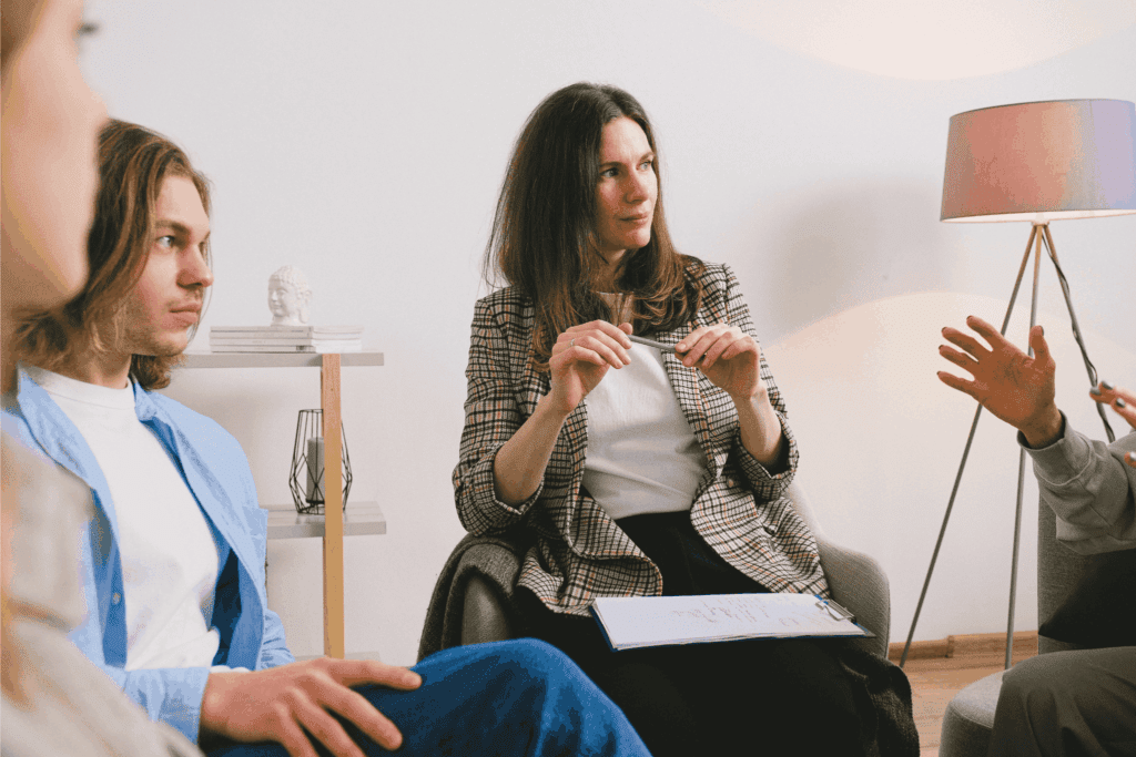 A small group engages in a therapy or counseling session, with one woman leading the discussion while others listen attentively.