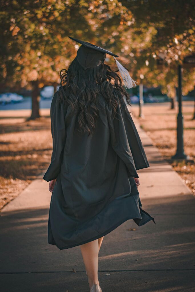 A graduate in cap and gown walks down a tree-lined path, celebrating their achievement.