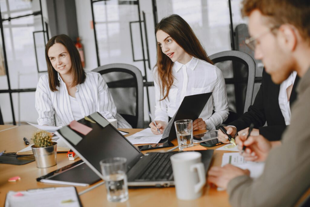 A group of colleagues sit around a conference table with laptops, notebooks, and documents, engaged in a business meeting or team discussion.