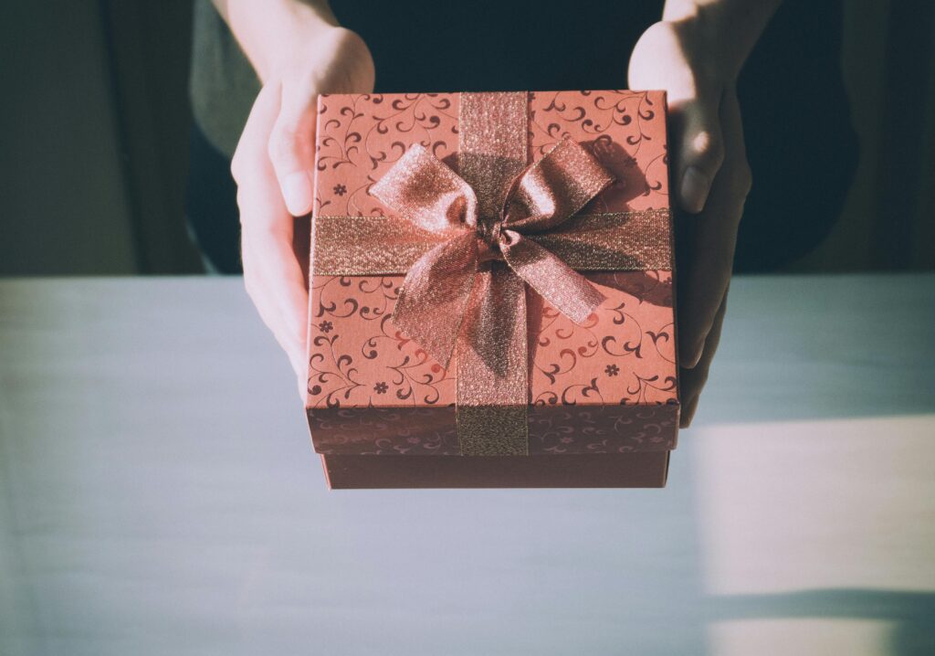 A person is holding out a beautifully wrapped gift box decorated with a patterned design and tied with a glittery ribbon bow.