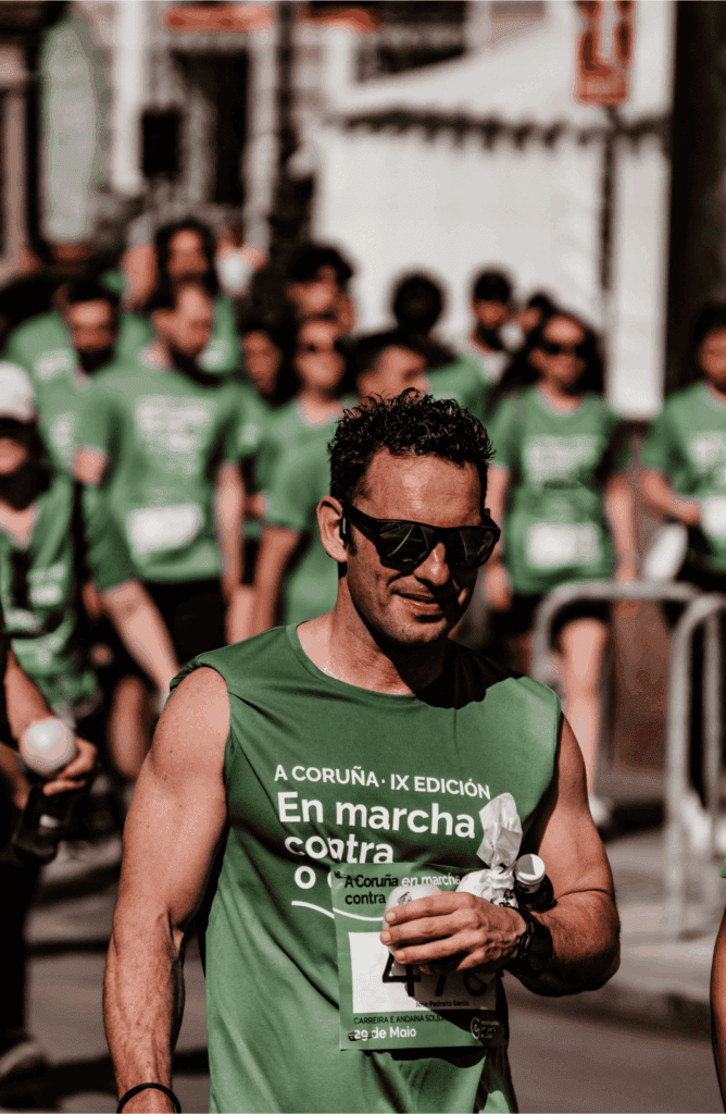 A man in sunglasses participates in a charity race, wearing a green shirt with other runners in matching shirts behind him.

