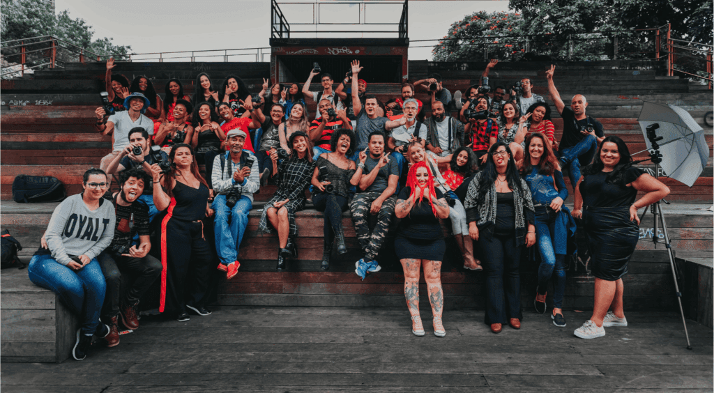 A large, joyful group poses together on wooden bleachers, with many people holding cameras and raising their hands in celebration.