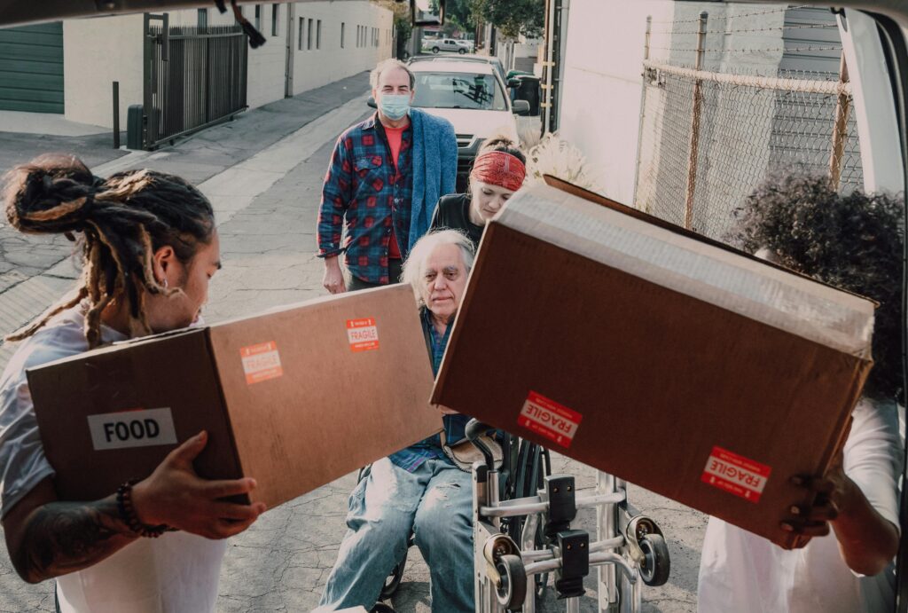 Volunteers delivering food and essential supplies to people in need, including an elderly man in a wheelchair.