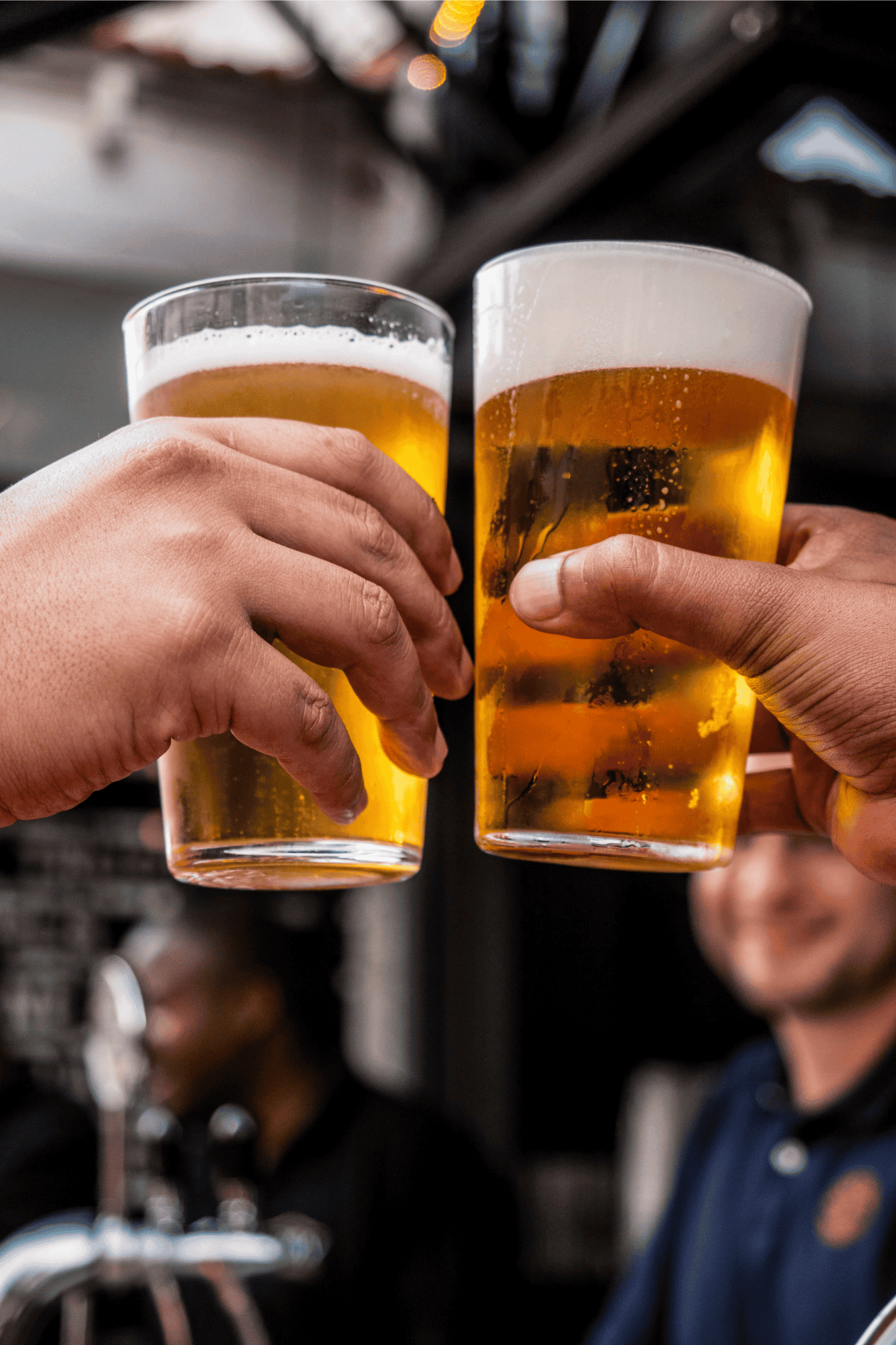 Two people clink glasses of cold, golden beer together in a celebratory toast at what appears to be a lively social gathering.