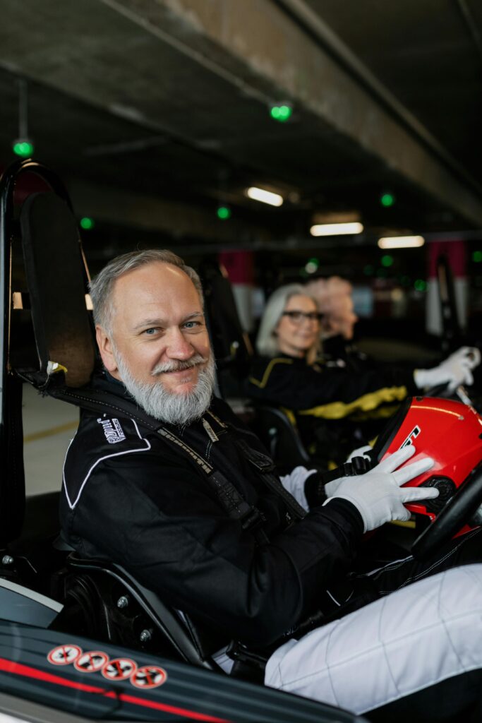 Older man in racing gear smiling while holding helmet in a go-kart