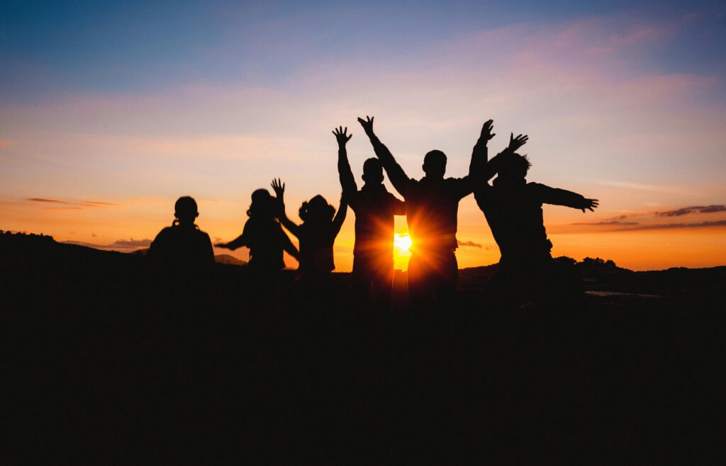 Team celebrating together with arms raised in front of a sunset, symbolizing teamwork and unity.