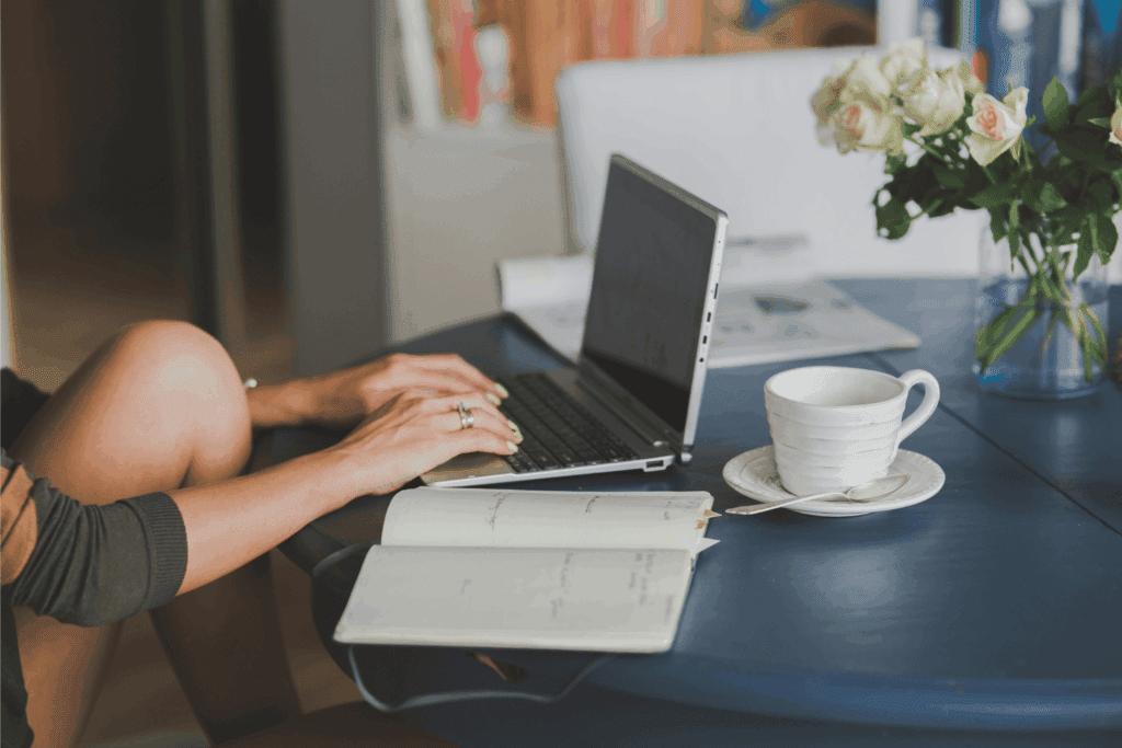 Person typing on laptop at a home office desk with an open notebook, coffee cup, and vase of white roses.