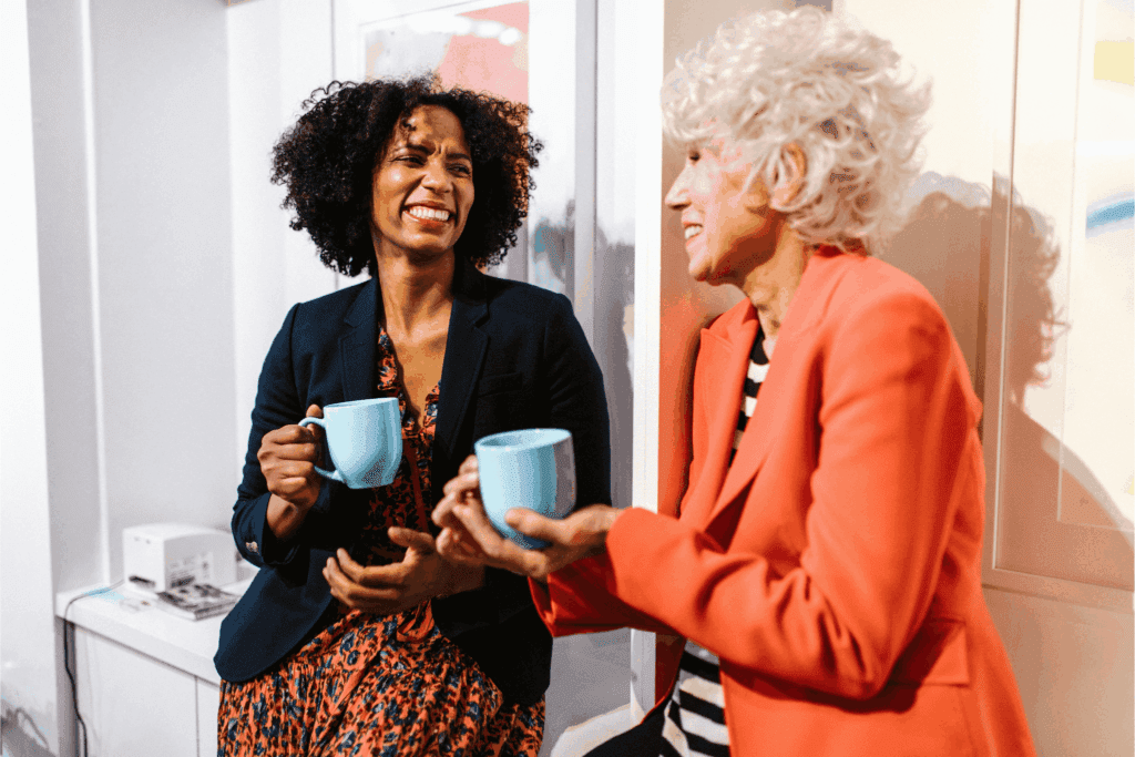 Two women smiling and enjoying a coffee break together while holding blue mugs in a bright office setting. 