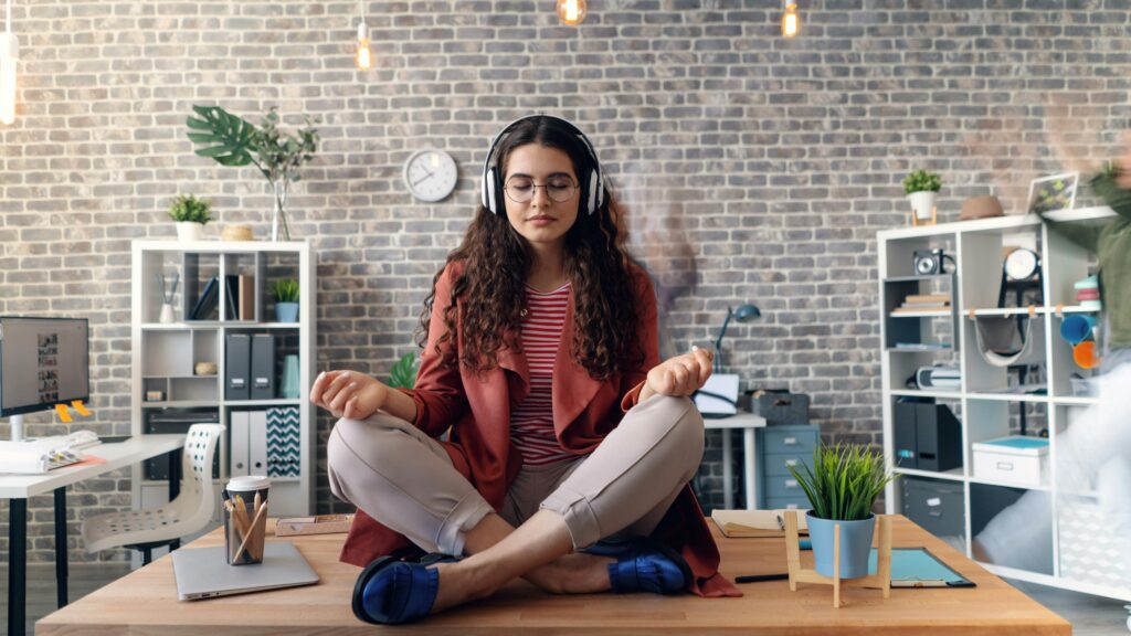 Young woman meditating with headphones on while sitting cross-legged on a desk in a modern office
