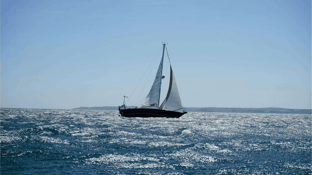 Sailboat gliding across sparkling blue ocean waves under a clear sky with distant coastline in the background