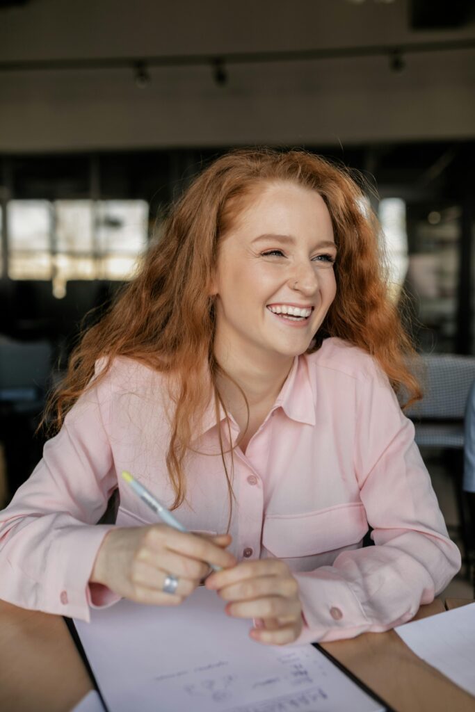 Smiling woman in a pink shirt holding a pen while working at a desk with papers