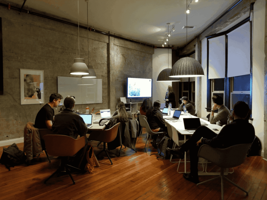 Team meeting in a modern office with employees working on laptops while a presenter leads a discussion using a screen.