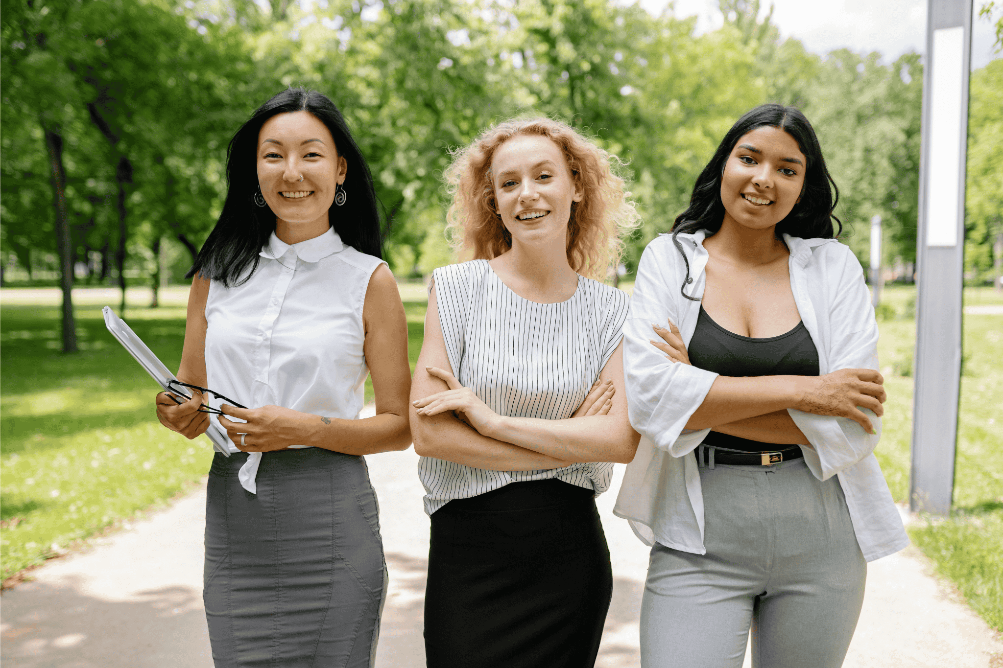 Three confident businesswomen standing outdoors in a park, smiling and posing together in professional attire together 