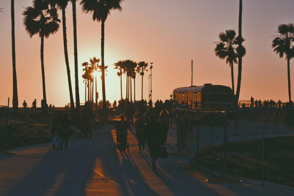 Sunset at Venice Beach Boardwalk with palm trees, people walking, and silhouettes against the ocean sky.